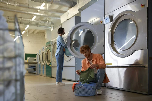 Young man holding clothes near basket and asian girlfriend in public coin laundry — Stock Photo