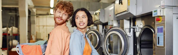 Усміхнений чоловік тримає кошик з одягом біля азіатської дівчини в монетному пральні, банер — стокове фото
