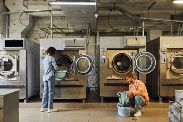 Mujer poniendo ropa en la lavadora cerca de novio con lavabo en la lavandería pública - foto de stock
