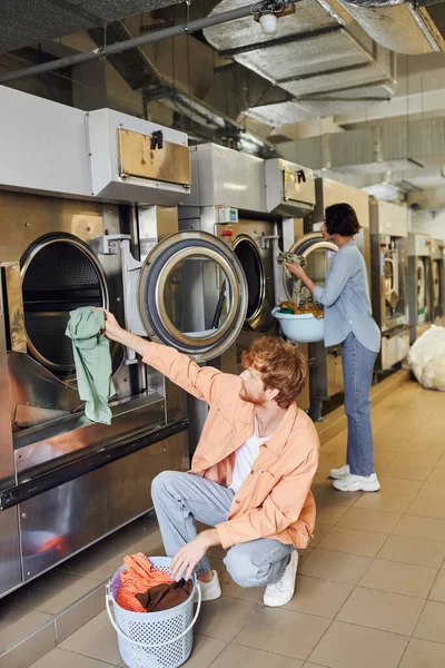 Человек кладет одежду в стиральную машину рядом с азиатской подругой в общественную прачечную монеты — стоковое фото