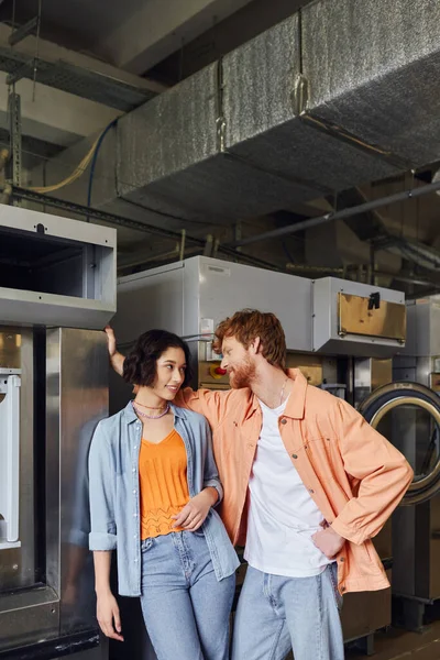 Молодой человек флиртует с азиатской подругой возле стиральных машин в публичной прачечной — стоковое фото