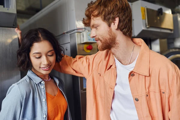 Young redhead man flirting with smiling asian girlfriend near washing machine in public laundry — Stock Photo