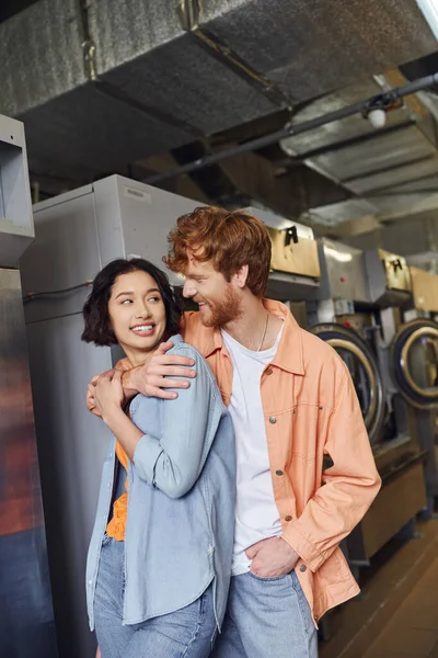 Sonriente joven pelirroja hombre abrazando asiático novia mientras de pie en público lavandería - foto de stock