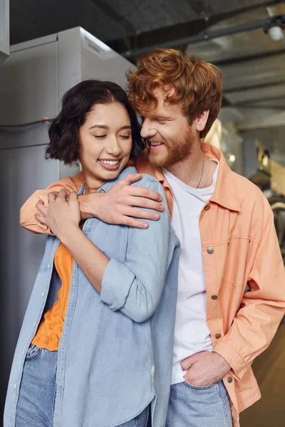 Gai rousse homme embrassant brunette asiatique copine tout flirt dans pièce blanchisserie — Photo de stock
