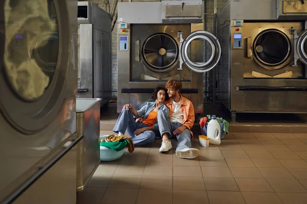 Giovane coppia romantica multietnica parlando mentre seduto vicino ai vestiti in lavanderia pubblica — Foto stock