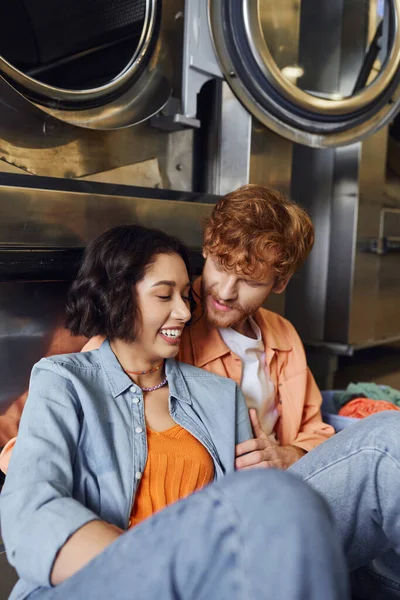 Ruiva homem abraçando alegre jovem asiático namorada perto de máquina de lavar roupa na lavanderia moeda — Fotografia de Stock