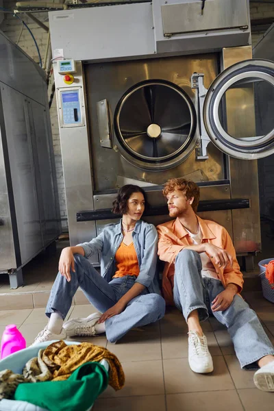 Giovane coppia interrazziale seduta vicino a vestiti e detergente sul pavimento in lavanderia pubblica — Foto stock