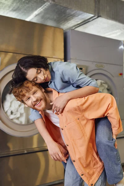 Young asian woman piggybacking on smiling boyfriend in blurred public laundry — Stock Photo