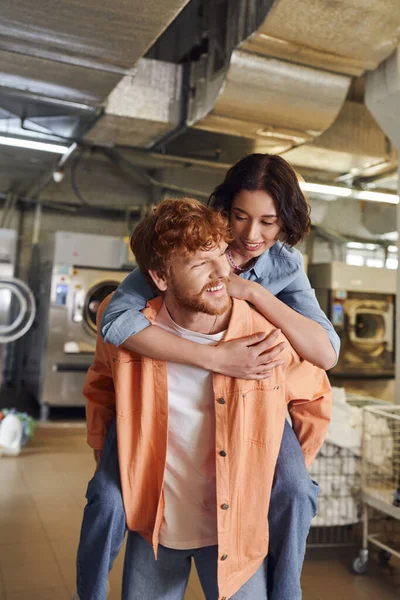 Cheerful young asian woman piggybacking on bearded boyfriend in blurred public laundry — Stock Photo