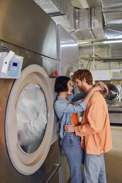 Vista lateral de una pareja interracial sonriente abrazándose mientras está de pie cerca de la lavadora en la lavandería de monedas - foto de stock