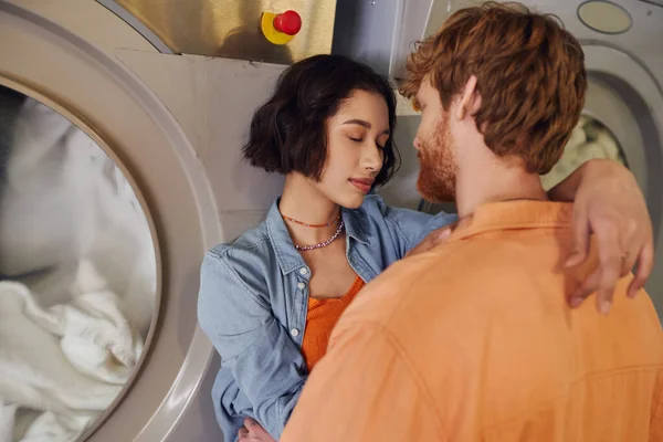 Romântico jovem asiático mulher abraçando ruiva namorado perto máquina de lavar roupa em público lavanderia — Fotografia de Stock