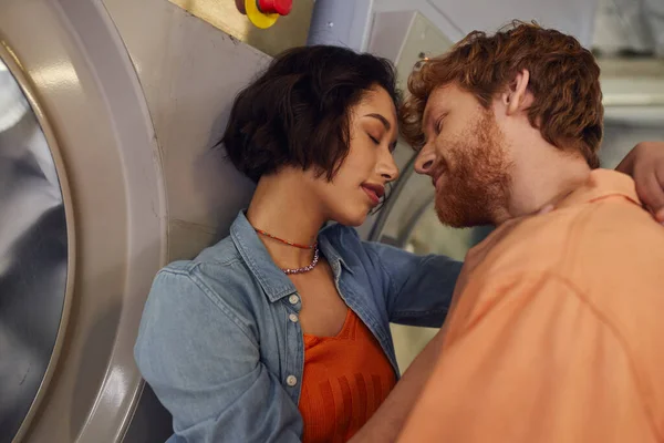 Jovem casal romântico multiétnico beijando perto de máquina de lavar roupa na lavanderia pública — Fotografia de Stock