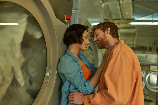 Sorrindo casal romântico multiétnico beijando perto de máquina de lavar roupa na lavanderia pública — Fotografia de Stock