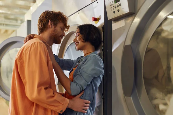 Alegre jovem casal interracial abraçando perto de máquina de lavar roupa na lavanderia pública — Fotografia de Stock
