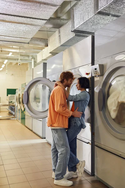 Vista lateral de jovens romântico casal multiétnico abraçando perto de máquinas de lavar roupa na lavanderia pública — Fotografia de Stock