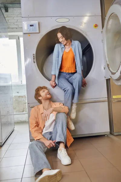 Alegre joven asiático mujer sentado en lavadora cerca pelirroja novio en público lavandería - foto de stock