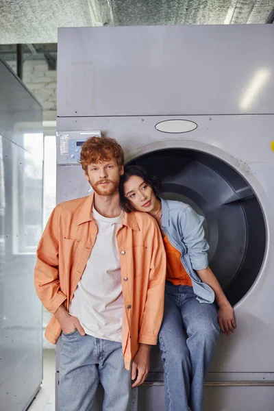 Young asian woman sitting on washing machine near boyfriend in public laundry — Stock Photo