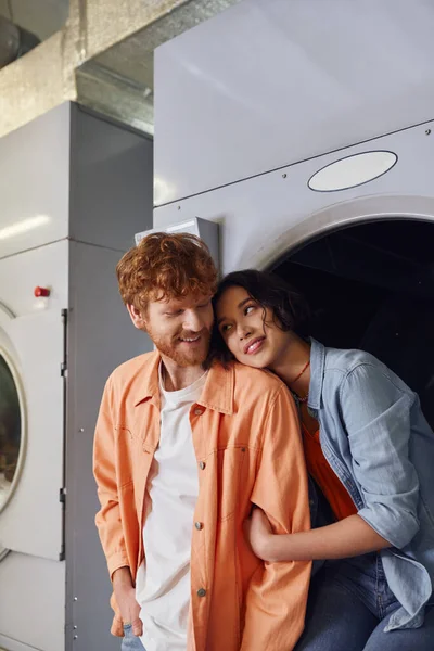Sorrindo jovem asiático mulher abraçando e olhando para namorado perto máquina de lavar roupa em público lavanderia — Fotografia de Stock