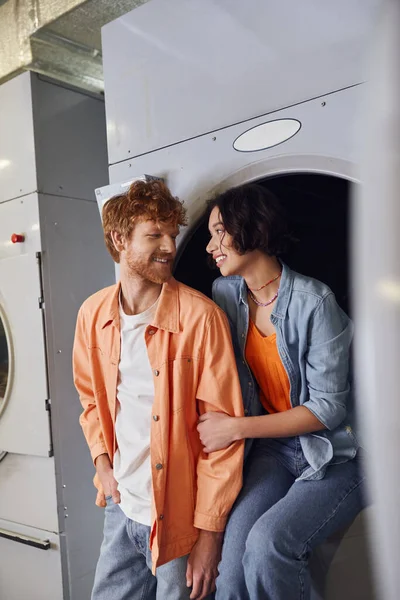 Cheerful young asian woman hugging redhead boyfriend while sitting on washing machine in laundry — Stock Photo