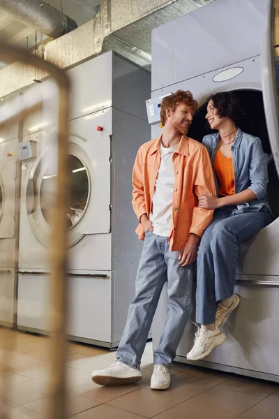 Comprimento total de alegre casal multiétnico falando perto da máquina de lavar roupa na lavanderia pública — Fotografia de Stock