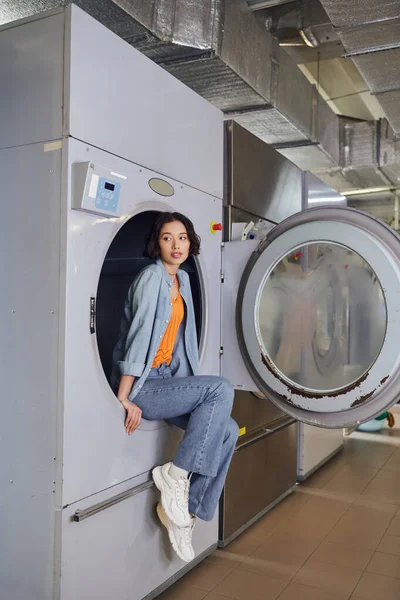 Full length of young asian woman sitting on washing machine in public laundry — Stock Photo