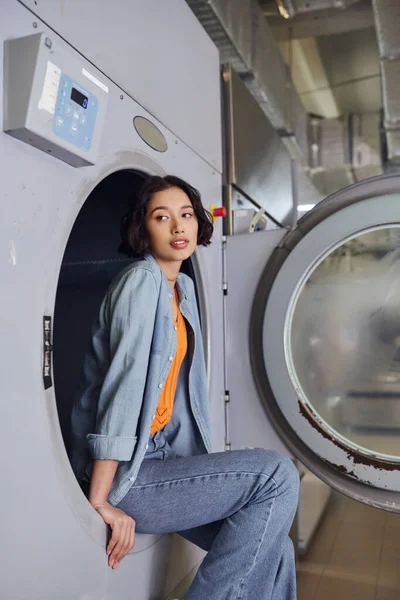 Jovem morena asiática mulher sentada na máquina de lavar roupa em lavanderia pública — Fotografia de Stock