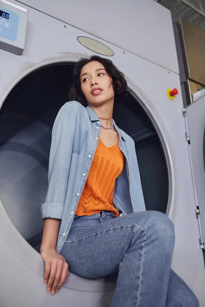 Baixo ângulo vista de jovem asiático mulher olhando para longe enquanto sentado na máquina de lavar roupa em público lavanderia — Fotografia de Stock