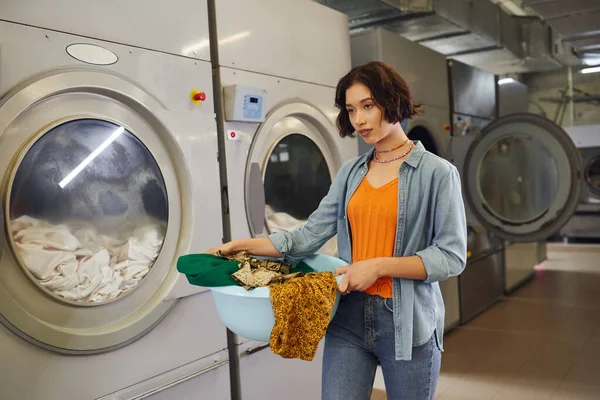 Young asian woman holding basket with clothes near washing machines in public laundry — Stock Photo