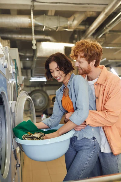 Smiling young man hugging asian girlfriend with clothes near washing machine in public laundry — Stock Photo