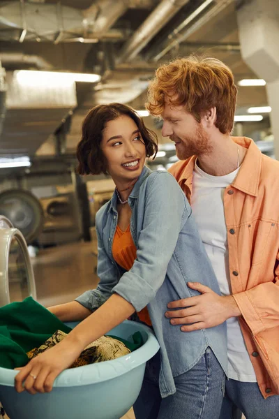 Rothaariger Mann umarmt fröhliche asiatische Freundin mit Kleidung in Waschbecken in der öffentlichen Wäscherei — Stockfoto
