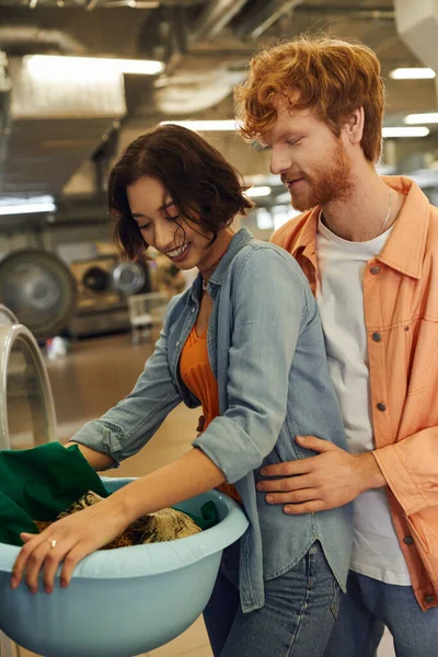 Redhead man hugging cheerful asian girlfriend with clothes and basket in coin laundry — Stock Photo