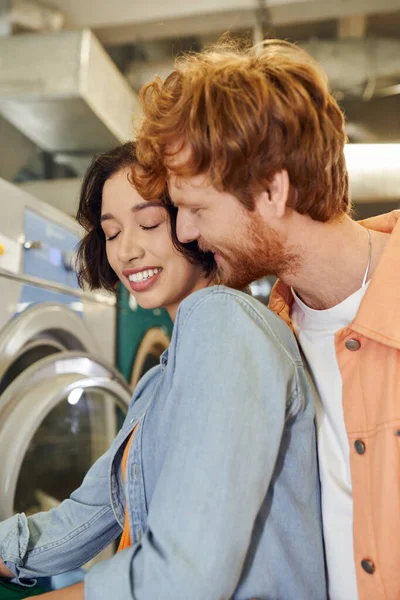 Young redhead man kissing cheerful asian girlfriend in blurred public laundry — Stock Photo