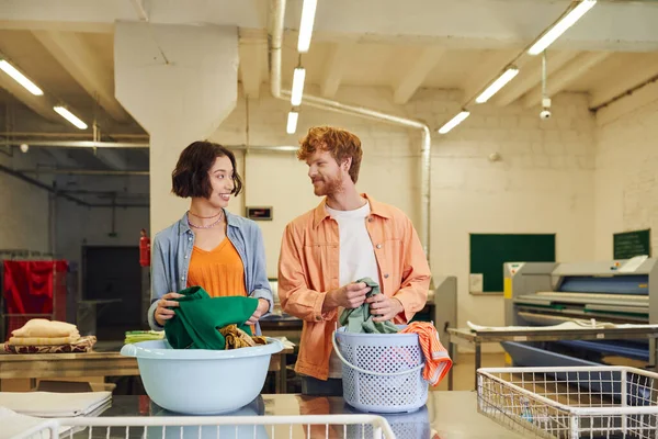 Sonriente interracial joven pareja hablando cerca de ropa y cestas en público lavandería - foto de stock