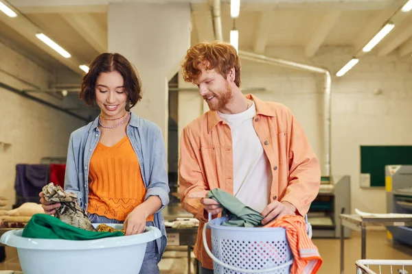 Lächelndes junges multiethnisches Paar, das Kleidung in Körbe in der öffentlichen Wäscherei auf dem Hintergrund legt — Stockfoto