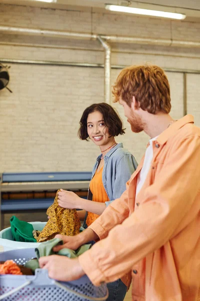 Cheerful young asian woman talking to boyfriend near clothes and baskets in coin laundry — Stock Photo