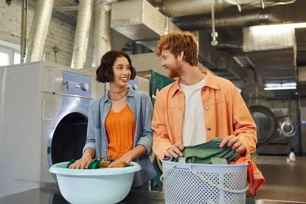 Laughing interracial couple talking near clothes in basins in public laundry on background — Stock Photo