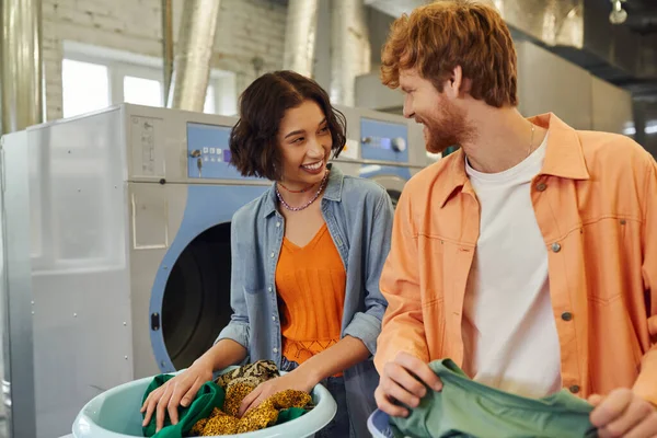 Joyful young asian woman talking to boyfriend while sorting clothes in public laundry — Stock Photo