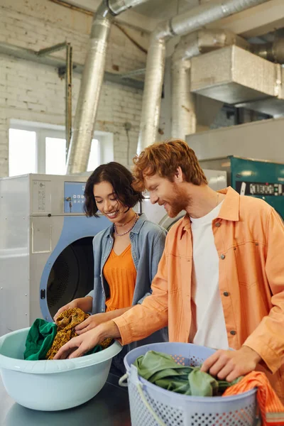 Despreocupados joven pareja multiétnica clasificación de ropa en cestas en la lavandería pública - foto de stock