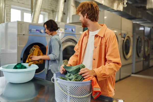Smiling man sorting clothes near basket and asian girlfriend in blurred public laundry — Stock Photo