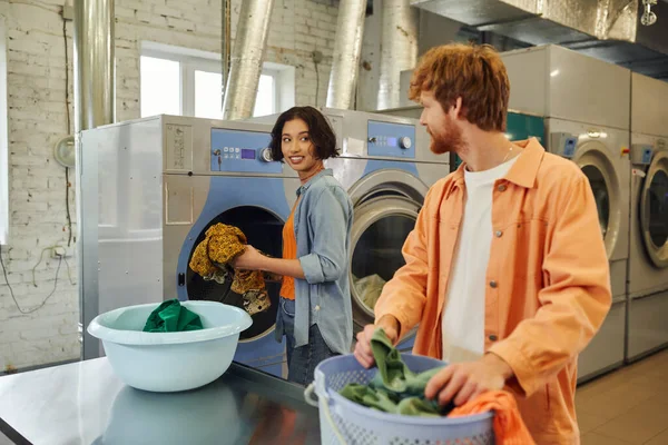 Smiling asian woman holding clothes near blurred boyfriend with basket in public laundry — Stock Photo