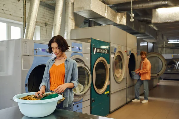 Joven asiático mujer clasificación ropa en lavabo cerca borrosa novio en público lavandería - foto de stock