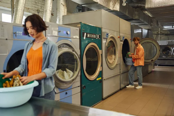 Young man standing near washing machine and blurred girlfriend with clothes in coin laundry — Stock Photo