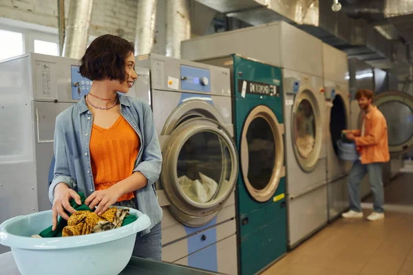 Smiling young asian woman looking at boyfriend near clothes in basin in blurred coin laundry — Stock Photo