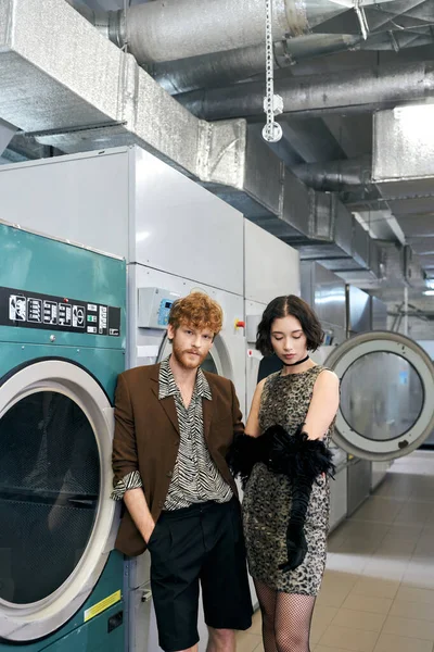 Elegante jovem casal multiétnico em pé perto de máquinas de lavar roupa na lavanderia pública — Fotografia de Stock