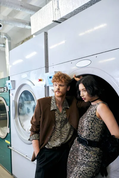 Уверенная в себе молодая многонациональная пара в стильных нарядах, стоящая возле стиральной машины в прачечной — Stock Photo