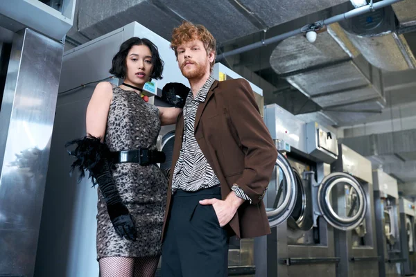 Baixo ângulo vista de moda jovem casal multiétnico olhando para a câmera na lavanderia pública — Fotografia de Stock