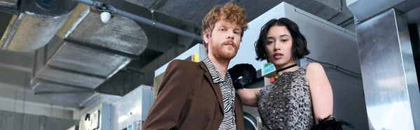 Vista de ángulo bajo de confianza joven pareja interracial mirando a la cámara en la lavandería de monedas, pancarta - foto de stock