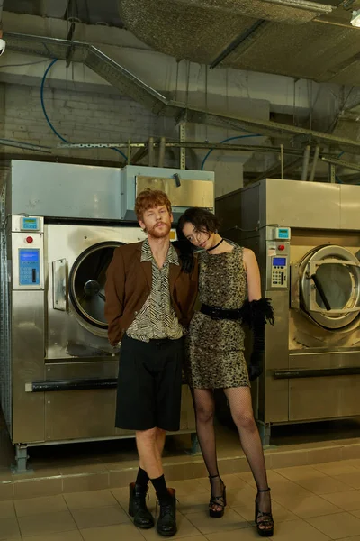 Comprimento total da moda jovem casal interracial posando perto de máquinas de lavar roupa na lavanderia pública — Fotografia de Stock