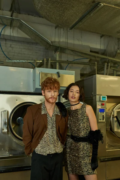 Retrato de moda joven pareja multiétnica mirando a la cámara en la lavandería pública por la noche - foto de stock