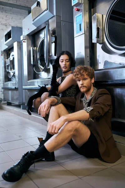 Confident man in jacket sitting near asian girlfriend in dress and washing machine in public laundry — Stock Photo
